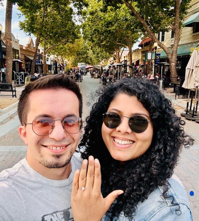 The couple taking a smiling selfie on a tree-lined downtown street, showing off the engagement ring